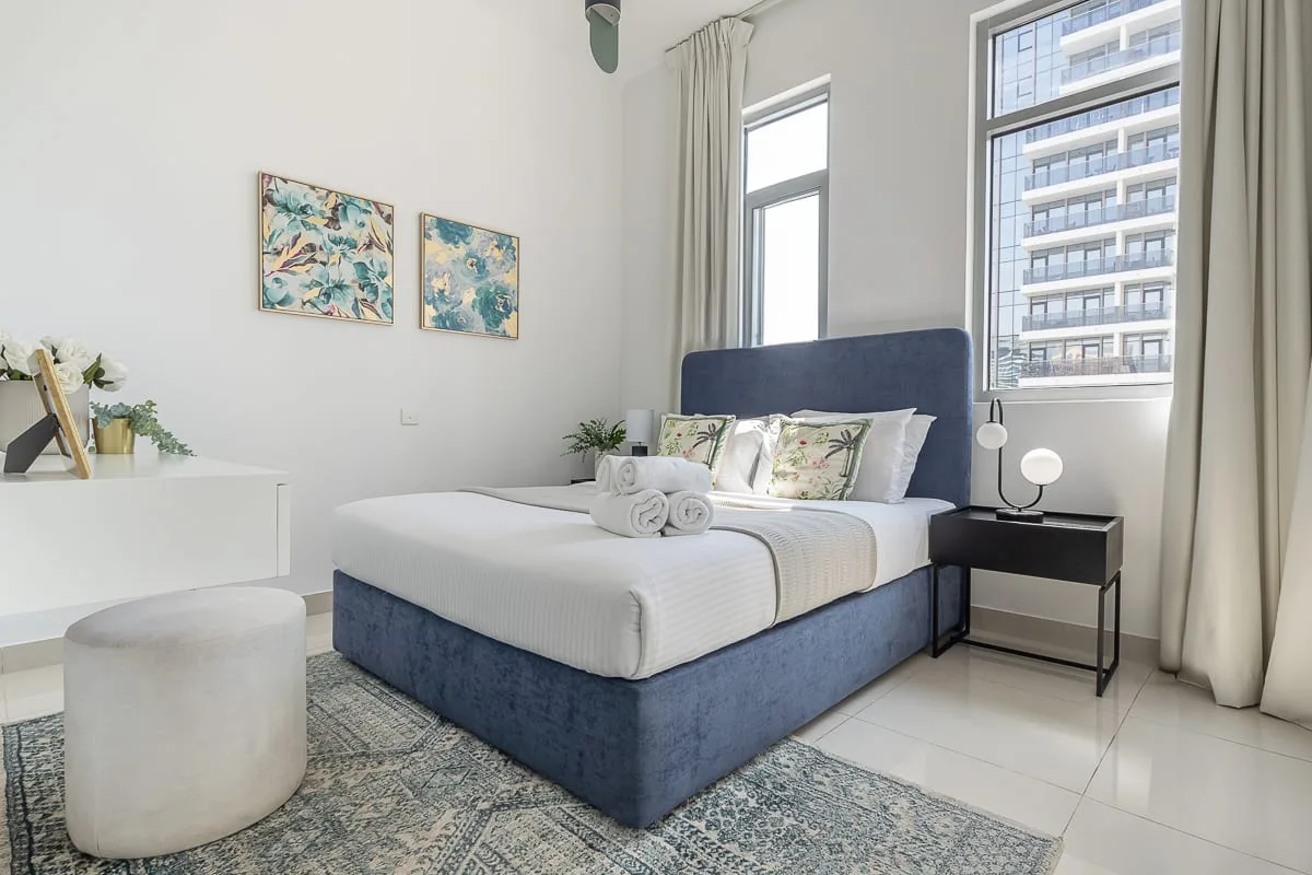 Un dormitorio luminoso con una cama junto a grandes ventanas y una pequeña mesa de noche.