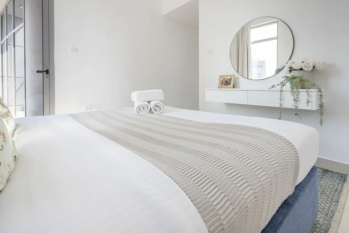 Un dormitorio luminoso con una cama bien tendida y un espejo redondo en la pared.