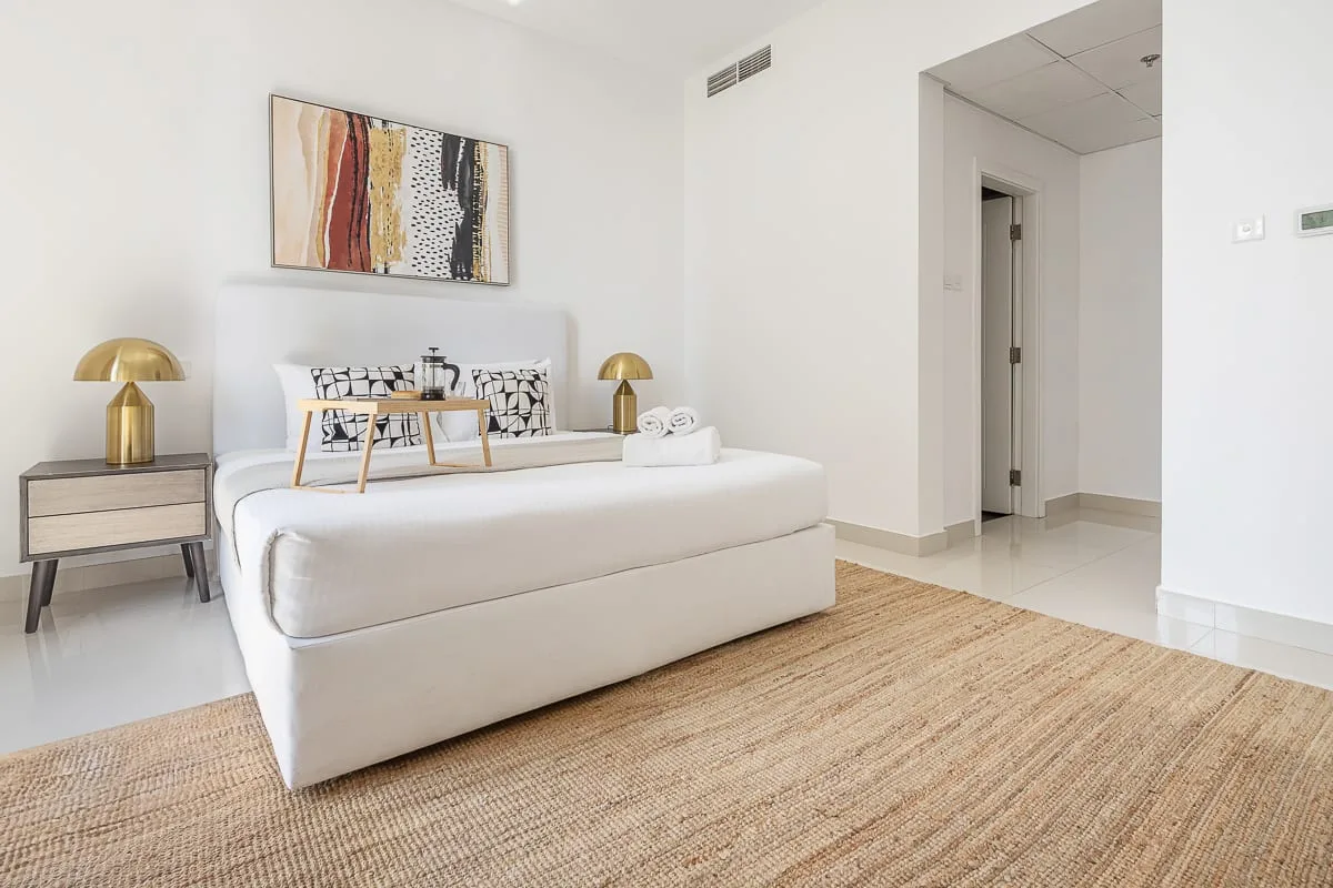 Un dormitorio luminoso con una cama blanca, mesitas de noche y una alfombra tejida.