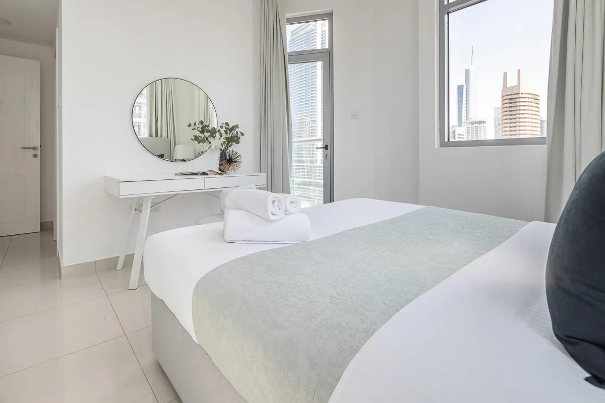 Un dormitorio luminoso con una cama arreglada y vistas a edificios cercanos.