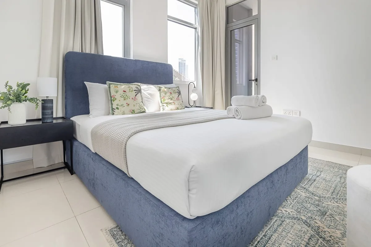 Un dormitorio con una cama bien tendida, cojines estampados y ventanas altas.