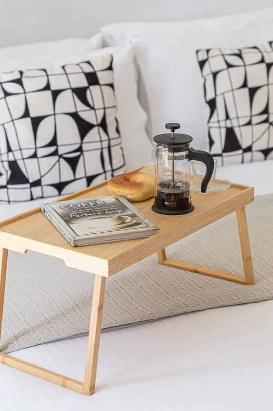 Una mesa bandeja de madera con café y un bollo está frente a un sofá blanco con cojines estampados.