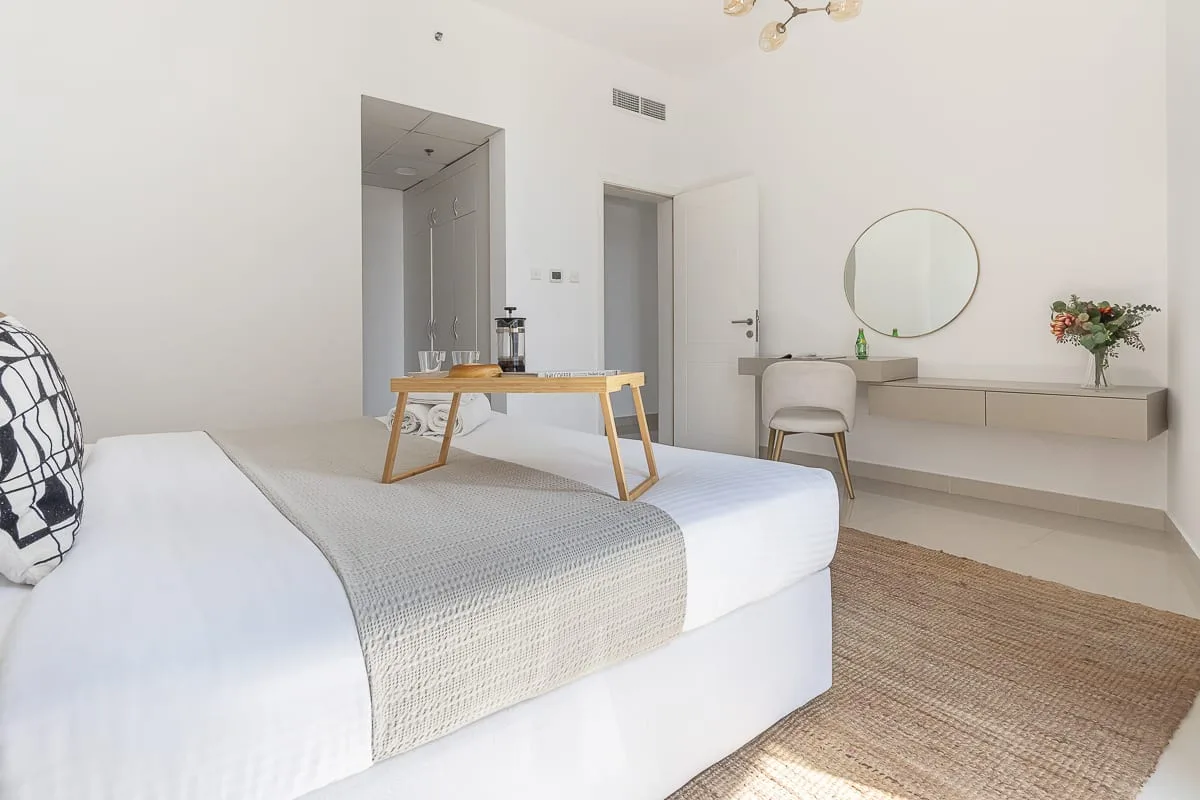 Un dormitorio luminoso con una cama bien tendida, una mesita y un tocador suspendido.