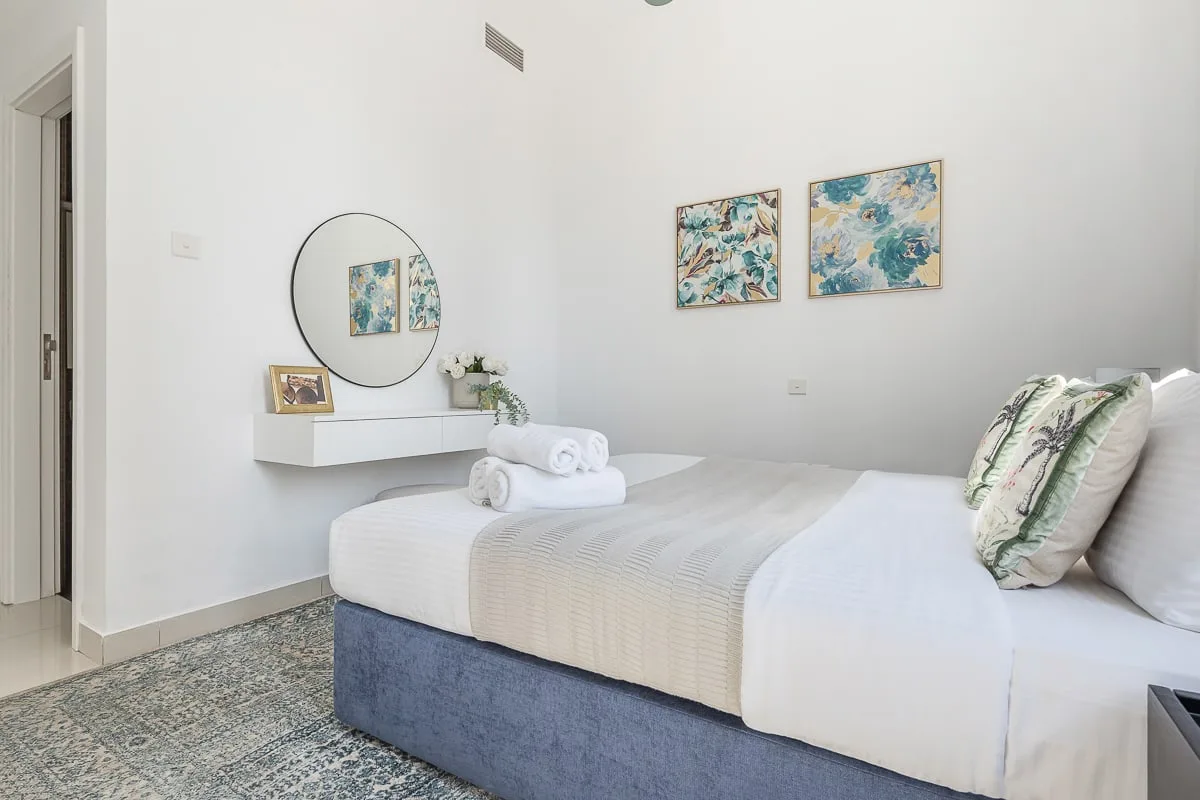 Un dormitorio luminoso con una cama bien hecha, cuadros coloridos y un espejo redondo.