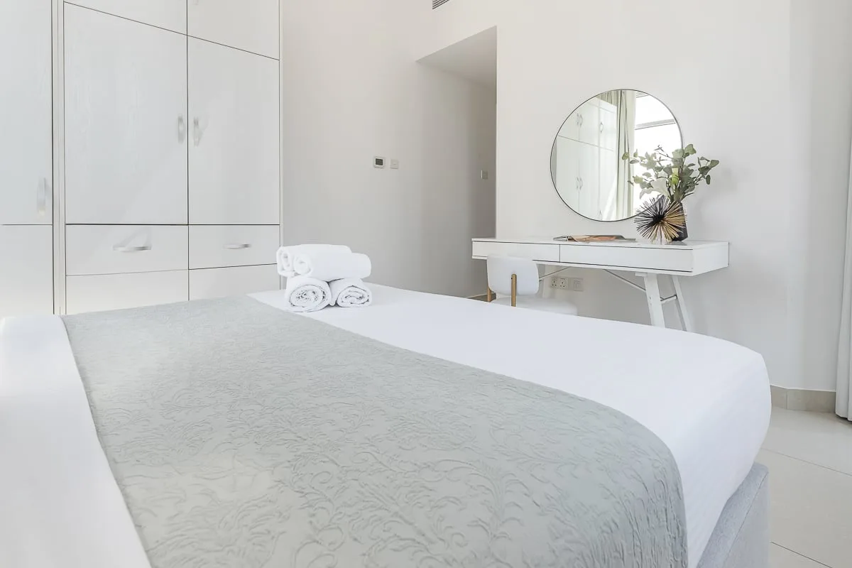 Un dormitorio luminoso con ropa de cama blanca y un tocador bajo un espejo redondo.