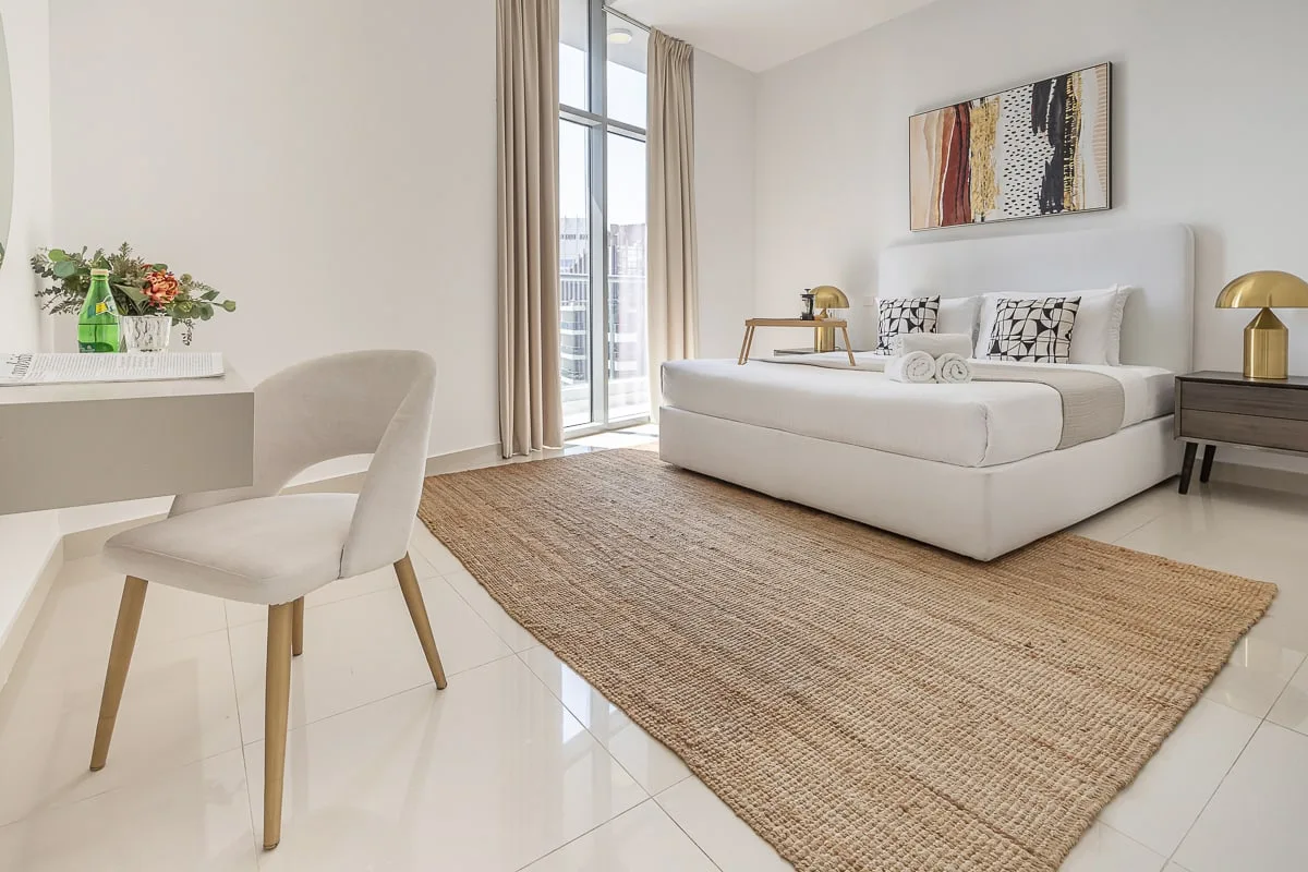 Un dormitorio luminoso con una cama, una alfombra tejida y ventanas altas.