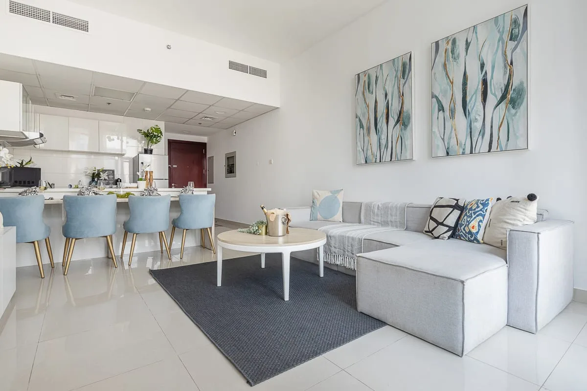 Una sala luminosa con cocina abierta, asientos claros y cuadros abstractos.