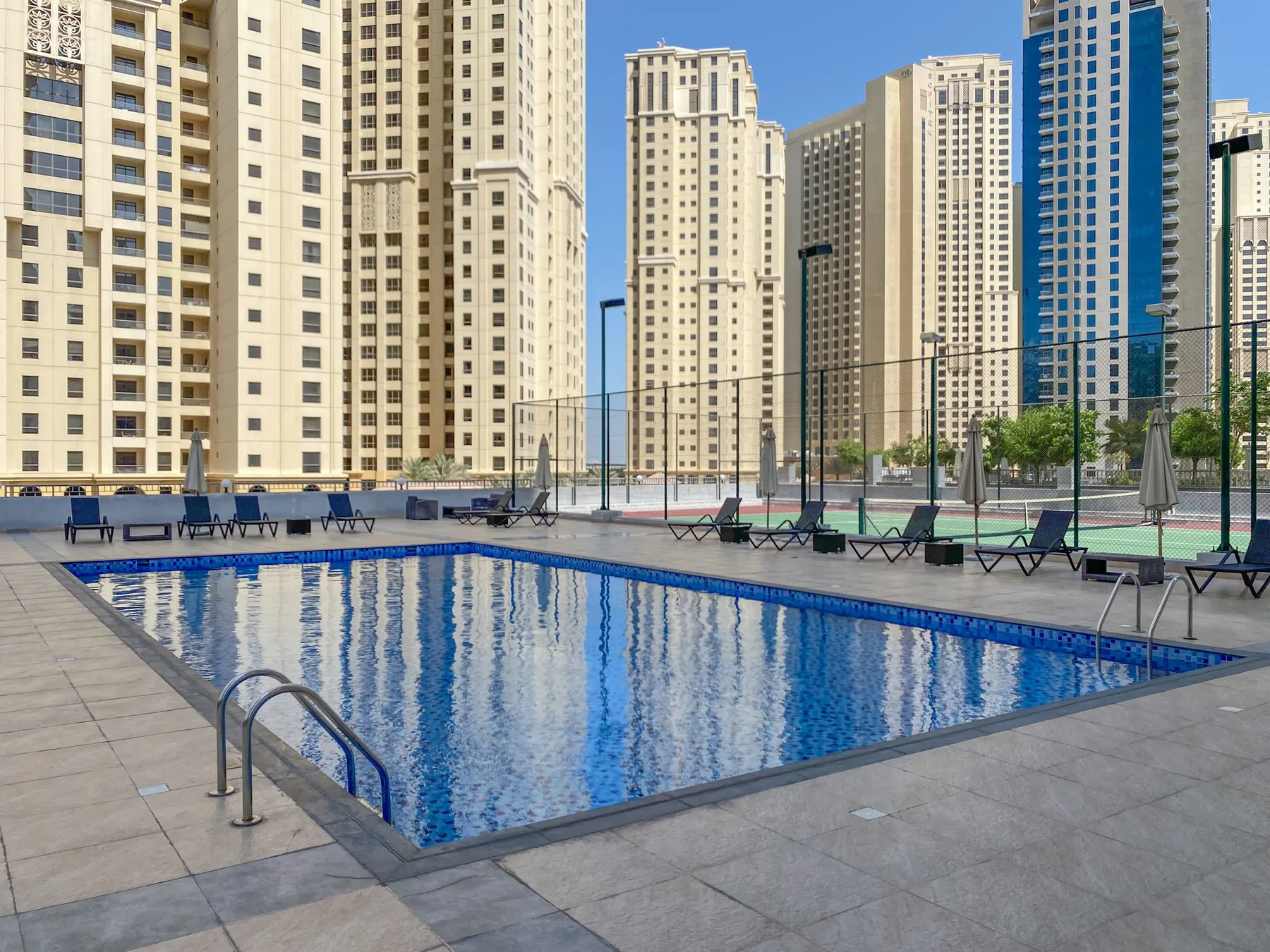Una piscina al aire libre está junto a una zona recreativa cercada y edificios altos.