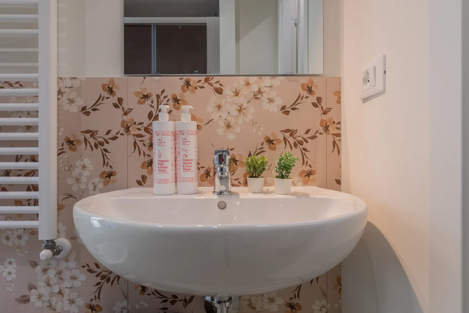 A bathroom sink stands against a floral-patterned wall with toiletries and small plants.
