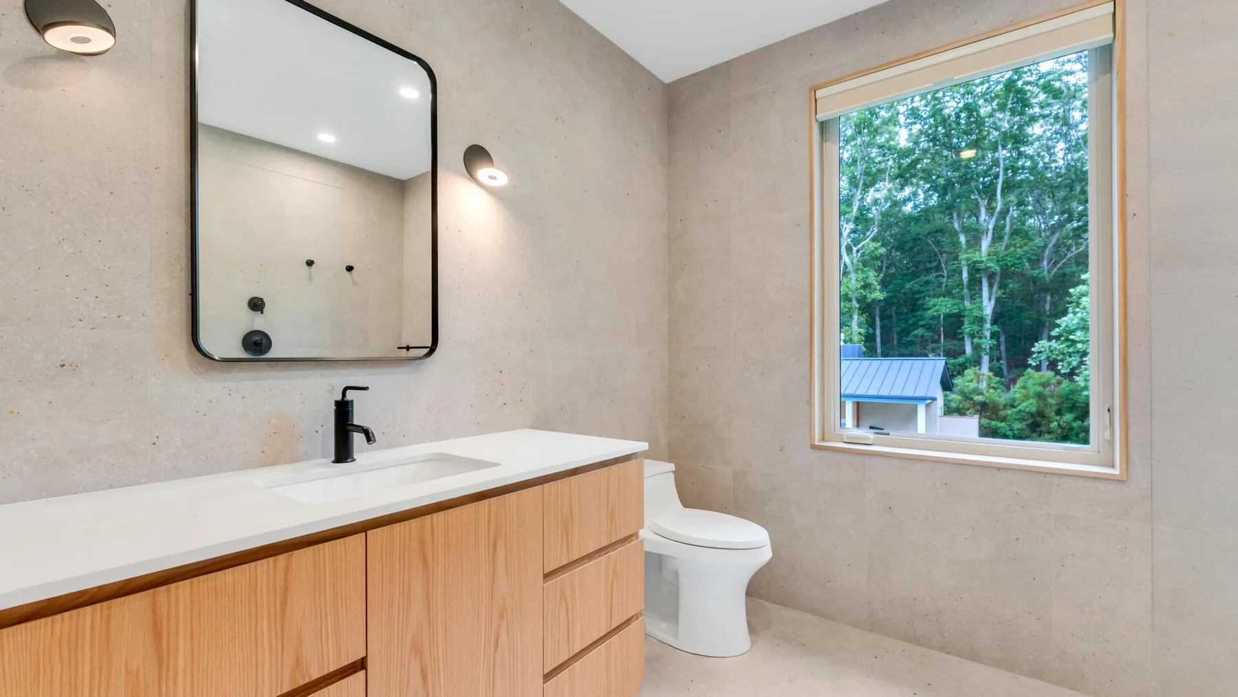 A bright bathroom with a long vanity, toilet, and window overlooking trees.
