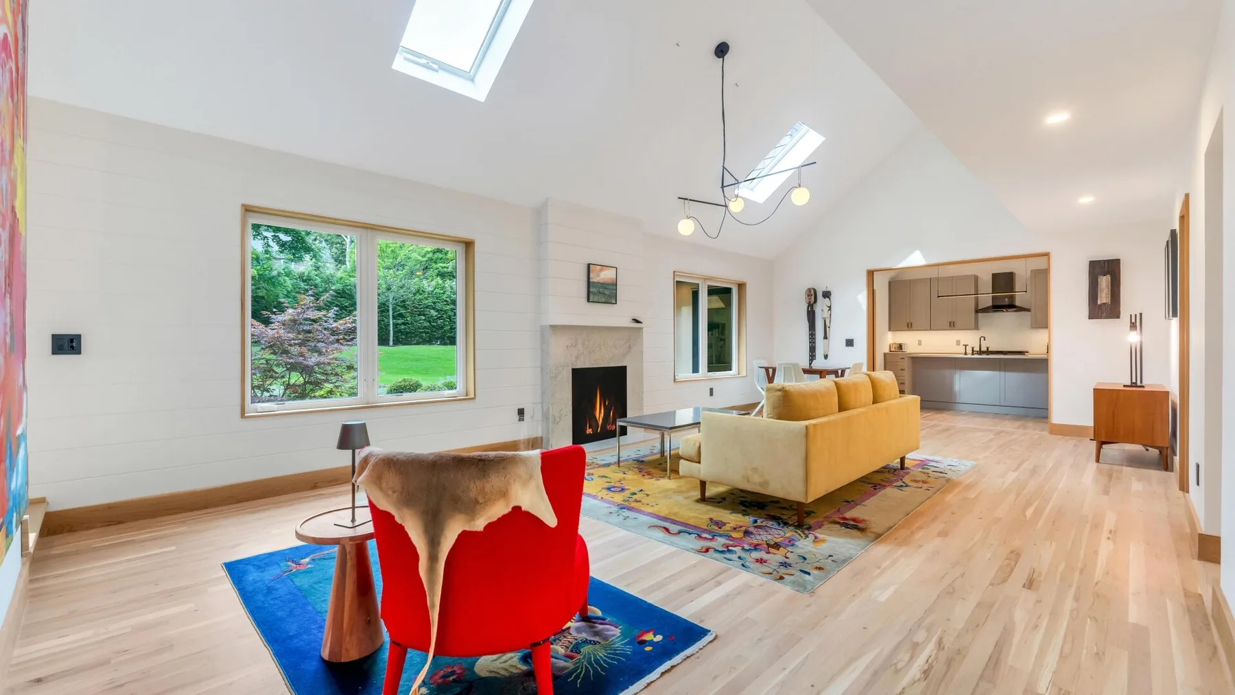 A bright open-plan living room with colorful seating, a fireplace, and a kitchen.
