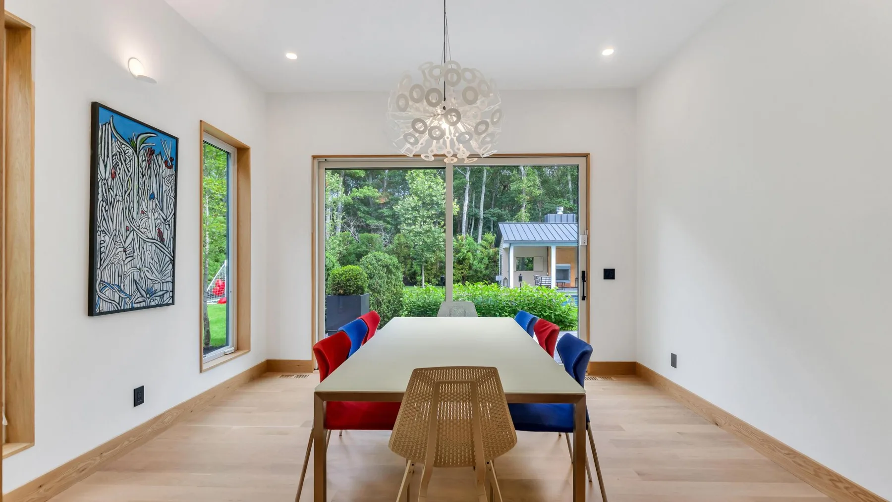 A bright dining room has a long table beside large windows overlooking greenery.