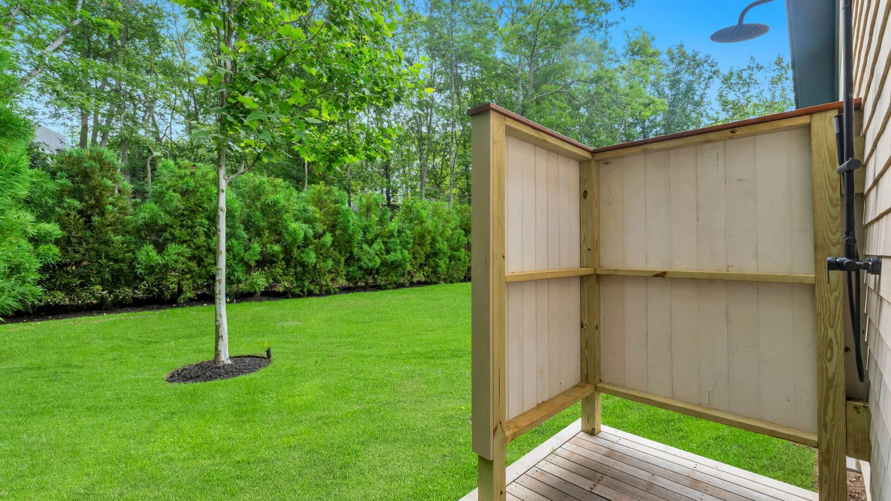 An outdoor shower area with a wooden privacy screen overlooks a green lawn and trees.