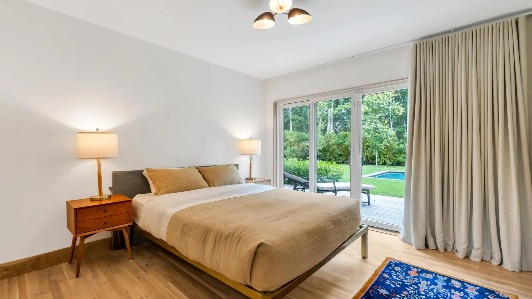 A bedroom with a bed beside large windows overlooking greenery and a pool.