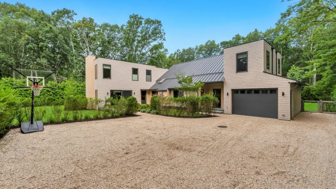 A modern house stands beside a broad gravel driveway and a basketball hoop.
