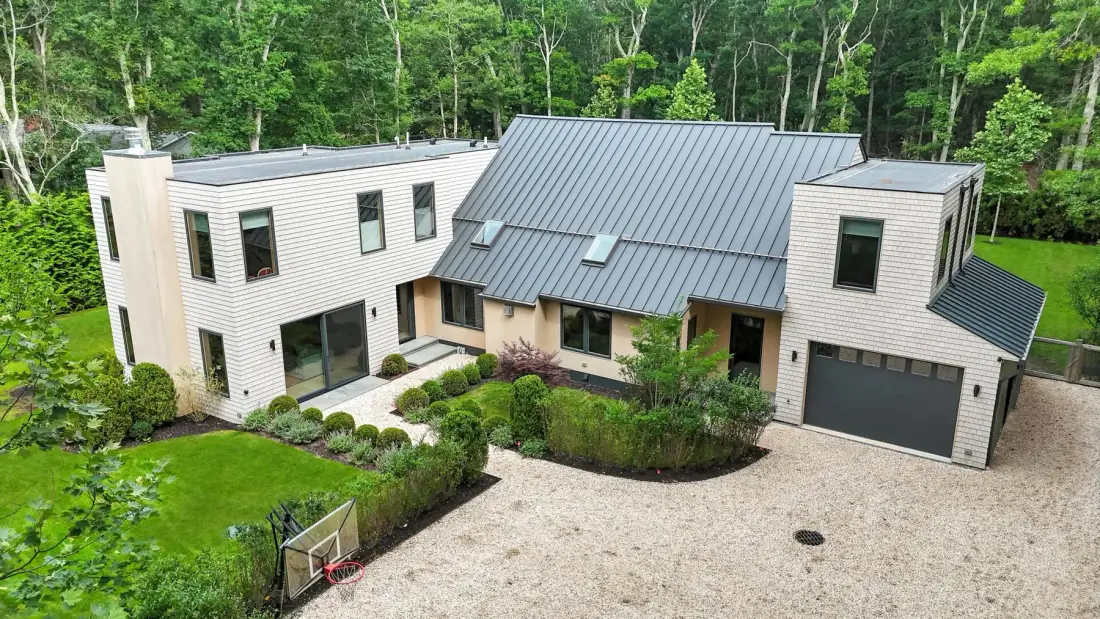 A modern house stands beside a gravel driveway, surrounded by trees and landscaping.