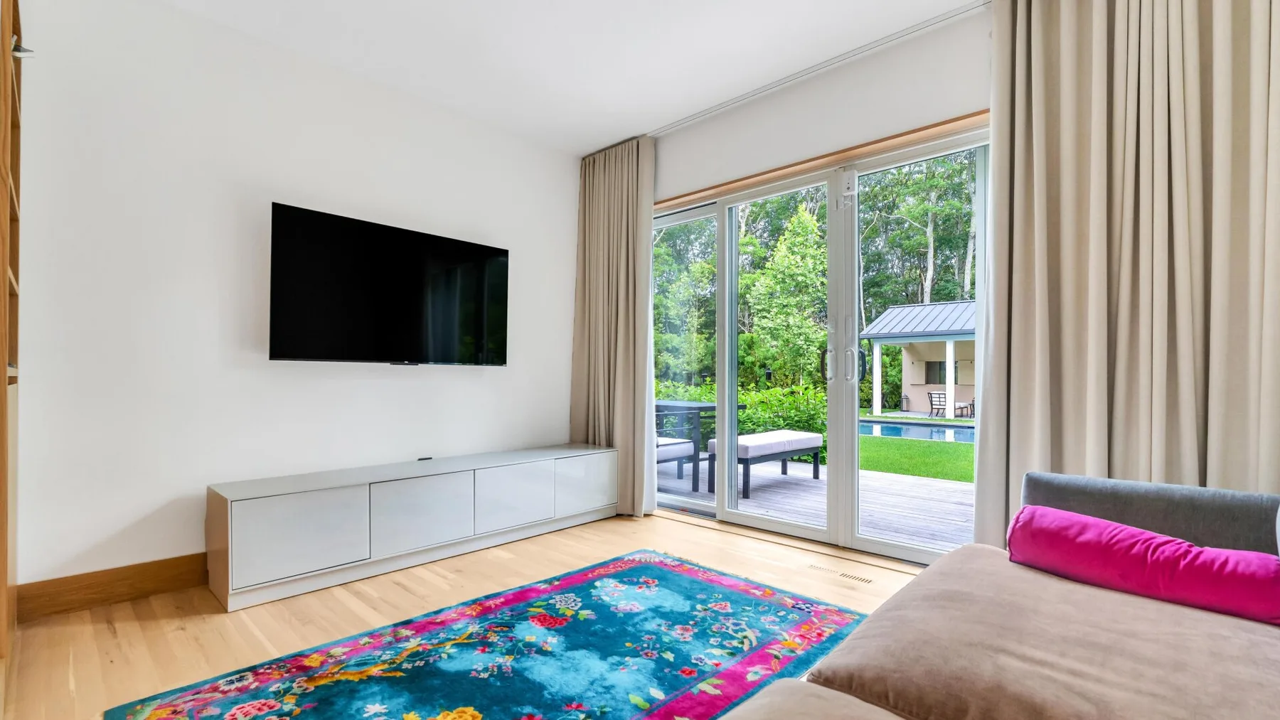 A bright living room with a wall-mounted television and large glass doors opening onto a deck.