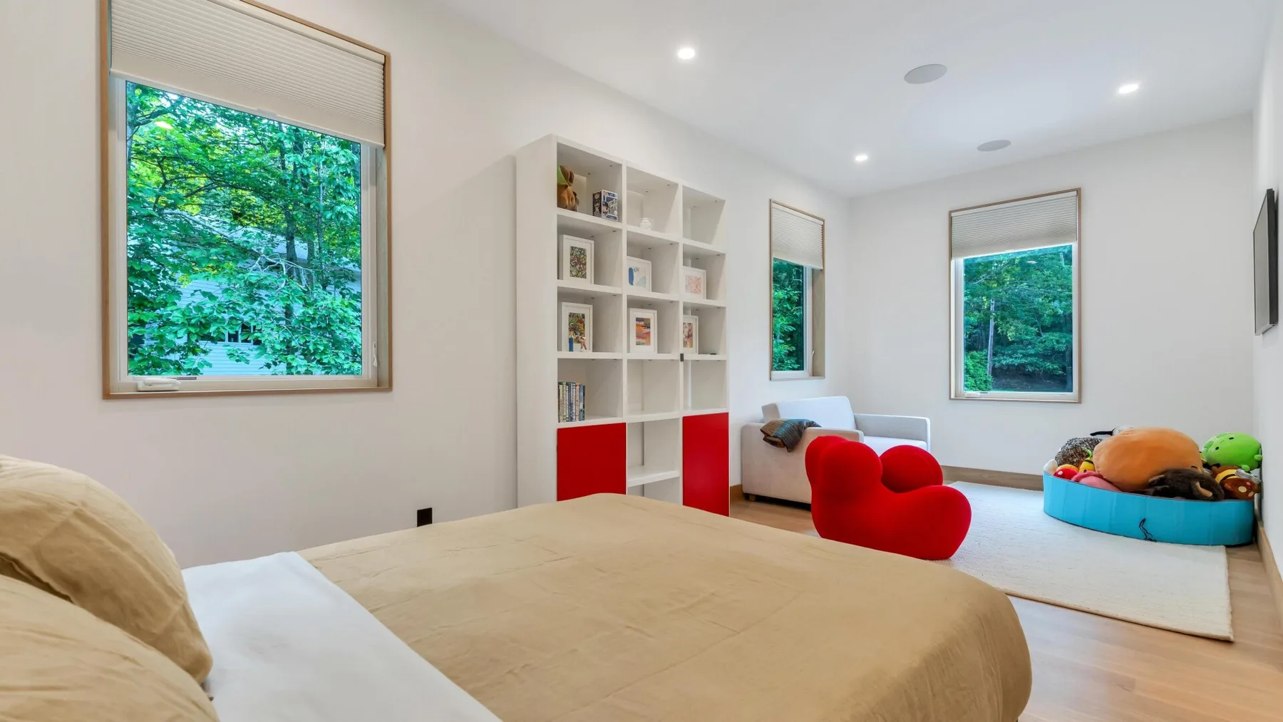 A bright bedroom with a large bed, a red chair, and windows overlooking greenery.