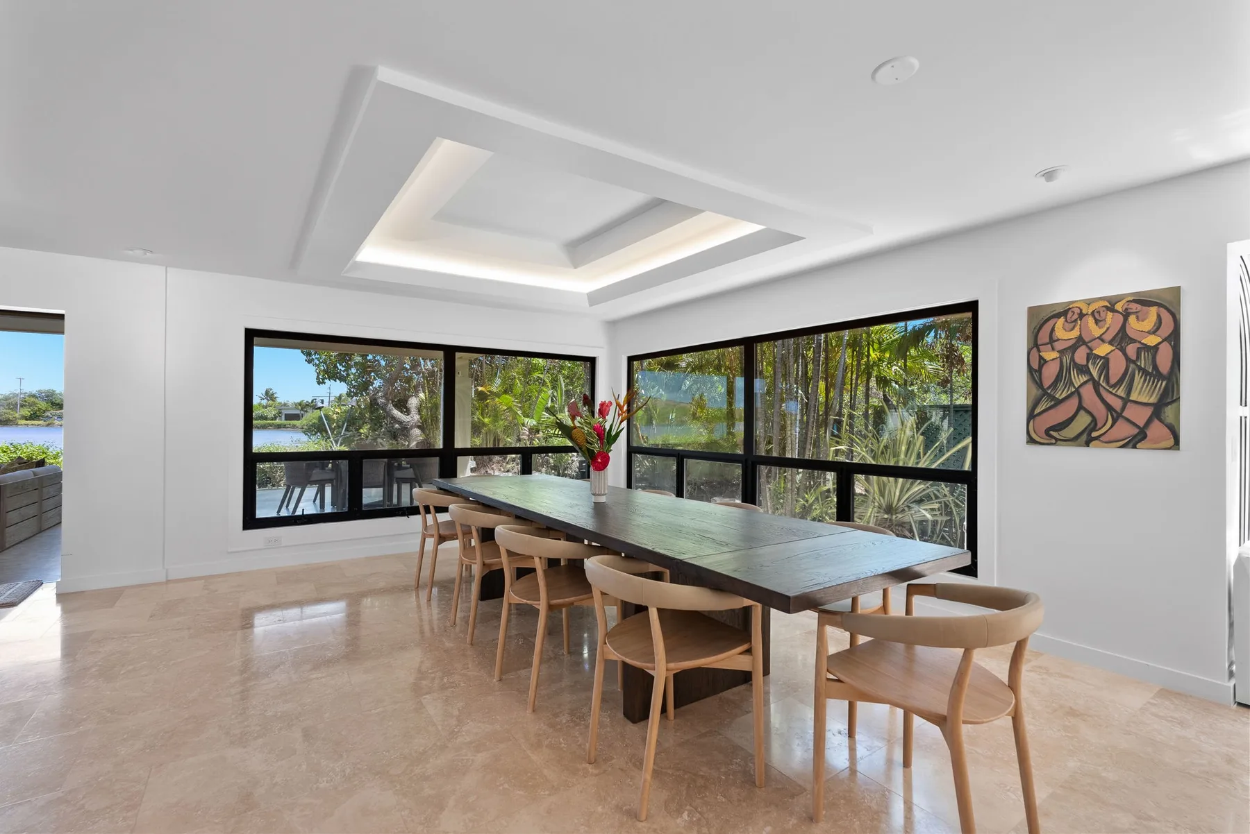 Un comedor luminoso tiene una mesa larga junto a grandes ventanas con vistas a árboles.