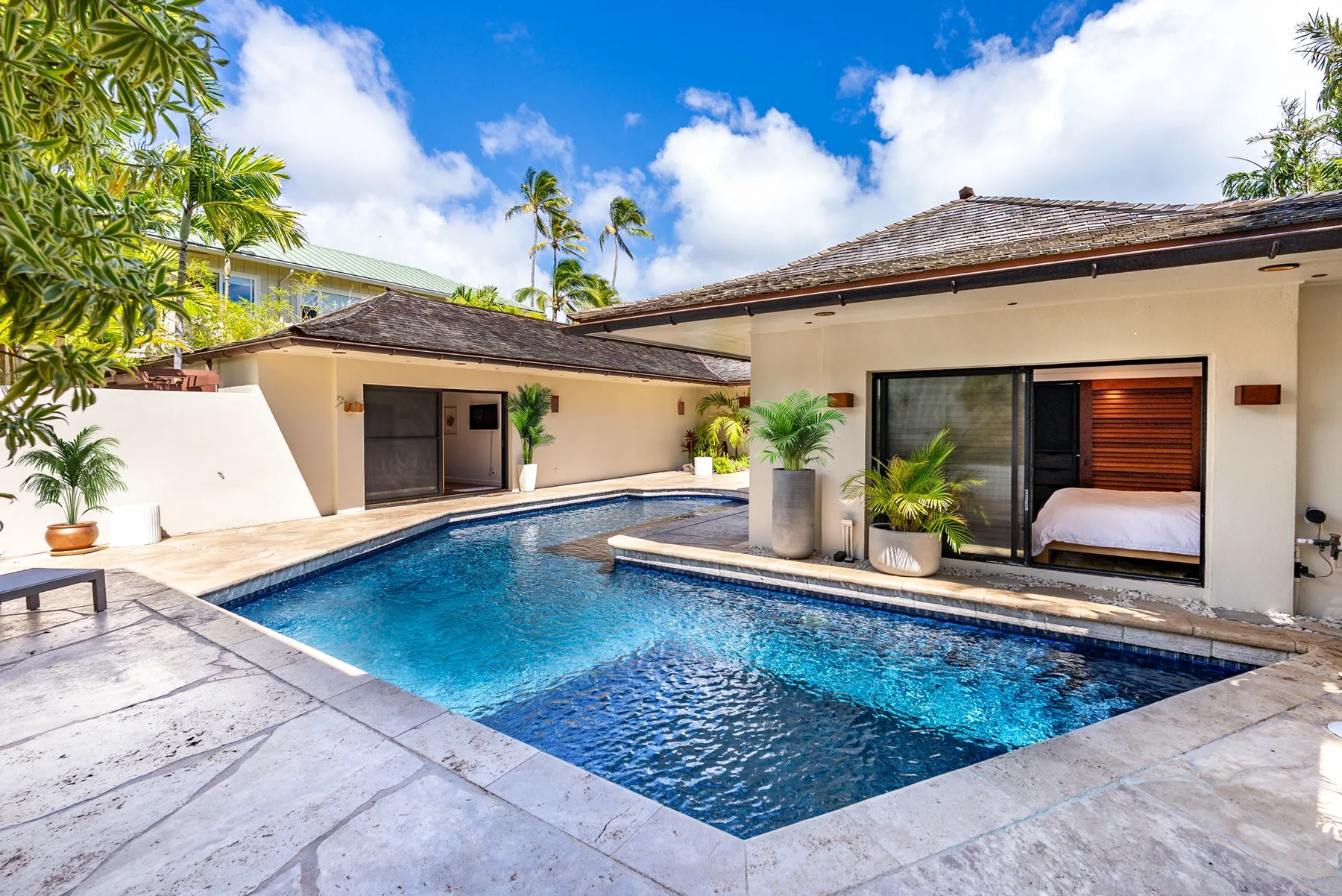 Una piscina junto a una vivienda de una planta, con patio cubierto y plantas tropicales.