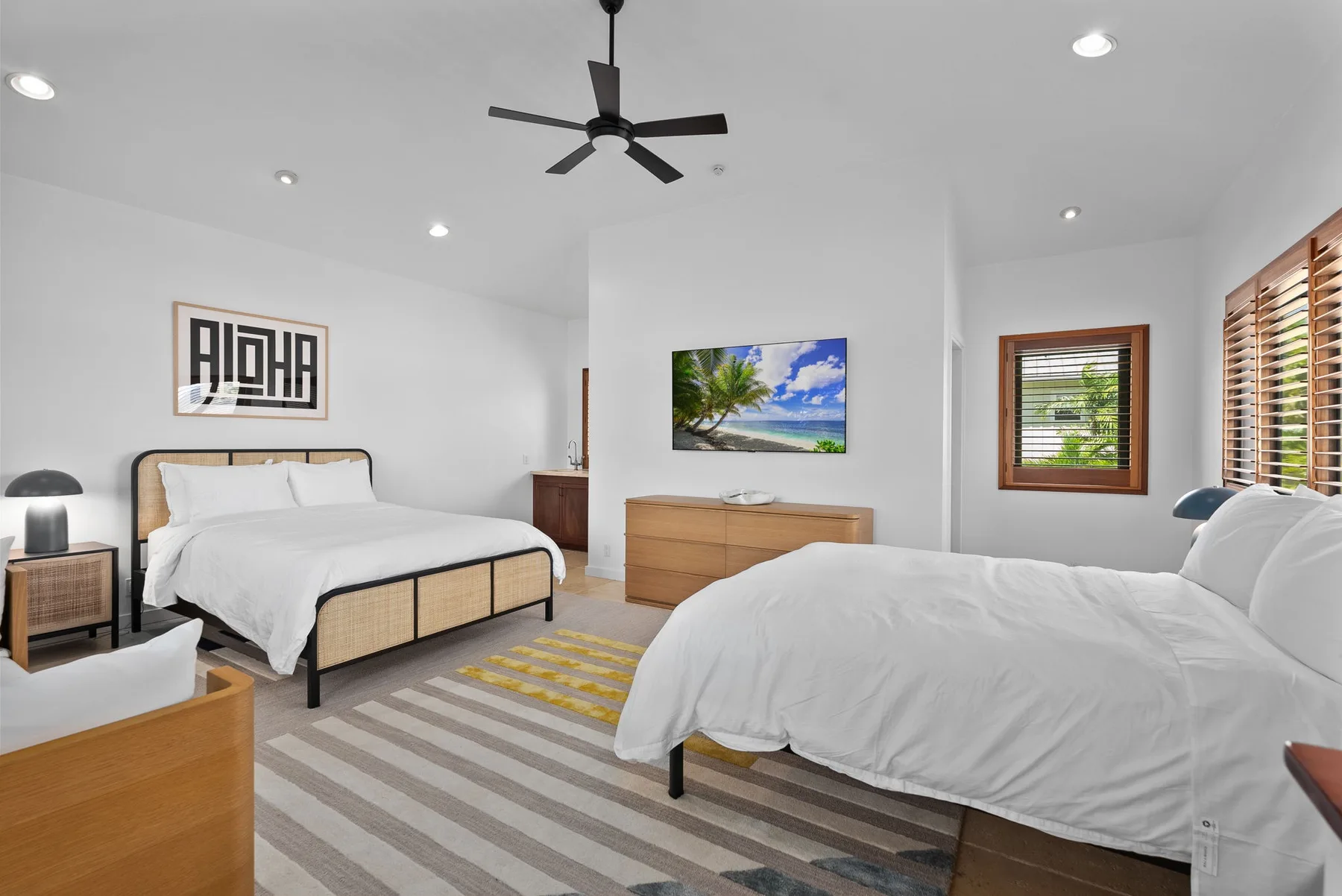 Un dormitorio luminoso con dos camas, un ventilador de techo y ventanas con persianas de madera.