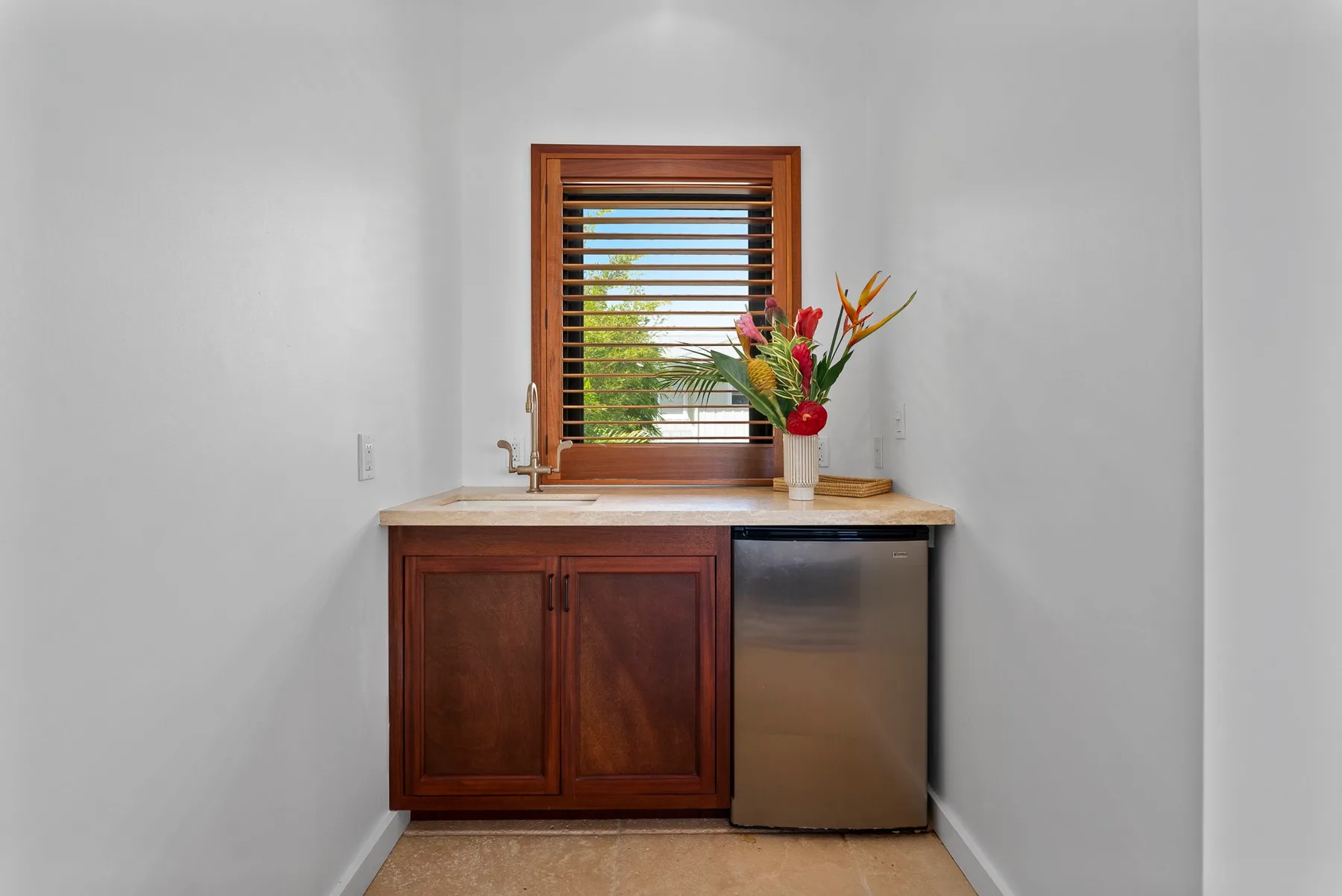Una encimera de cocina compacta está bajo una persiana de madera, junto a un pequeño arreglo floral.