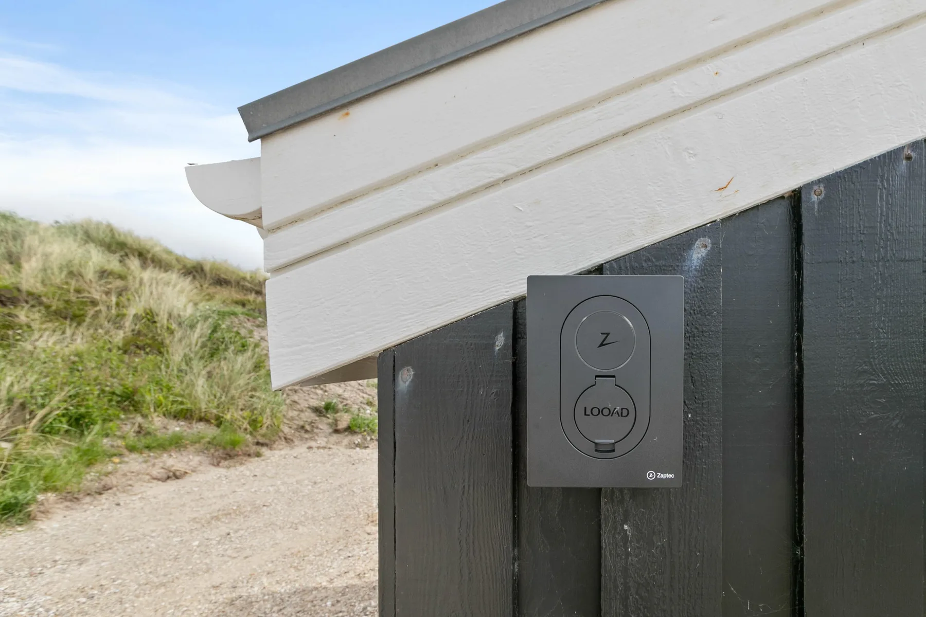 Una pared exterior oscura junto a un sendero arenoso, con un pequeño panel de control.