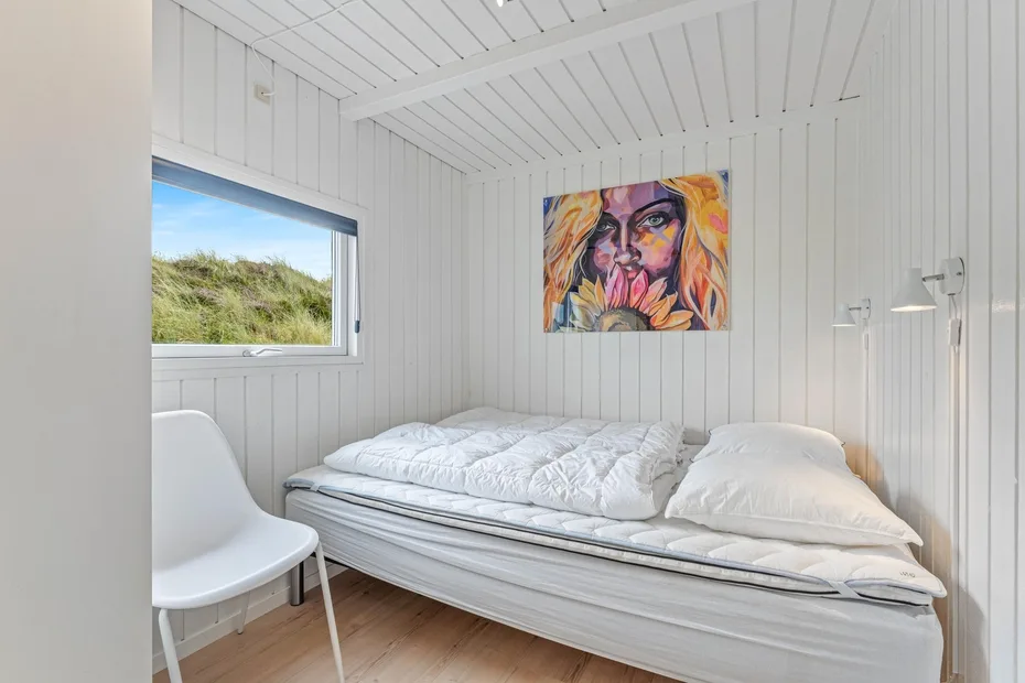 Un dormitorio luminoso con una cama, paredes blancas paneladas y un retrato colorido encima.