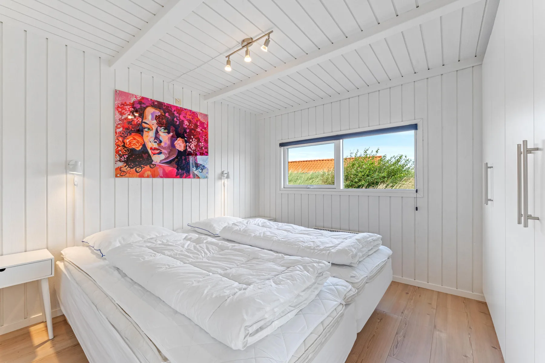 Un dormitorio luminoso con ropa de cama blanca y una obra colorida en la pared.