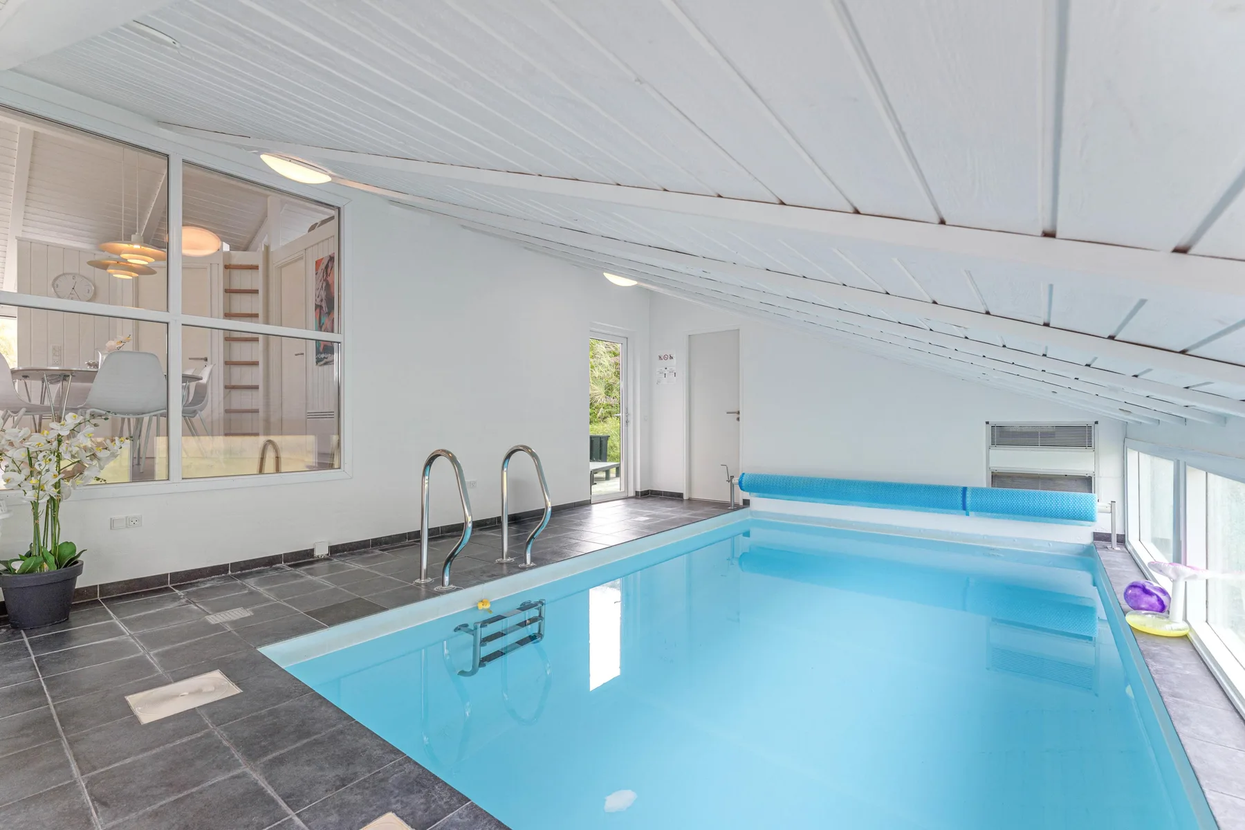 Una piscina cubierta está junto a grandes ventanas y una terraza de baldosas.