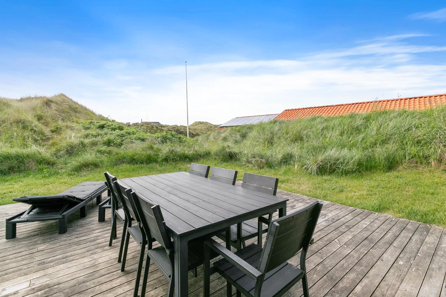 Una mesa de comedor y asientos al aire libre están en una terraza de madera entre dunas cubiertas de hierba.