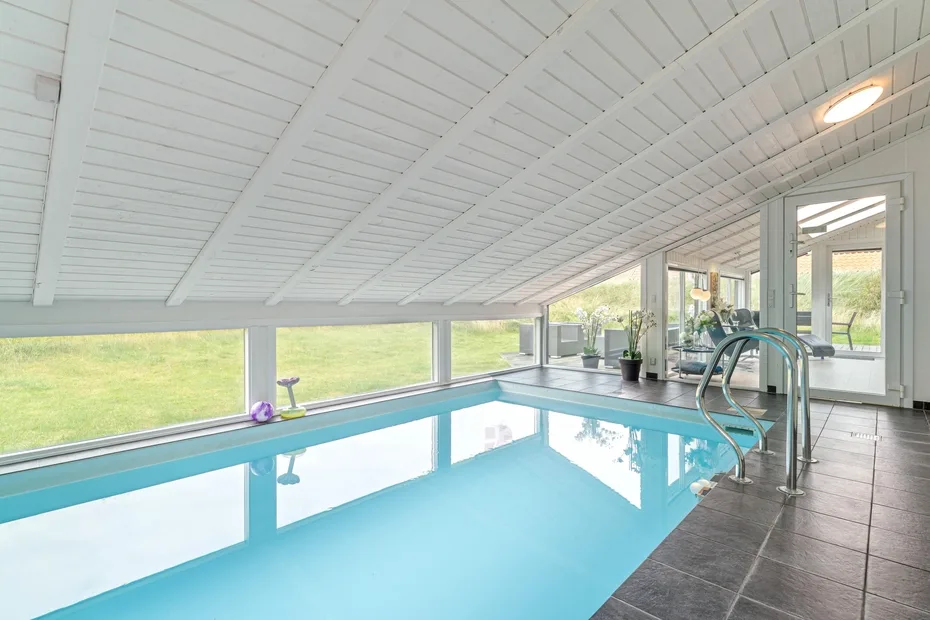 Una piscina cubierta se encuentra bajo un techo blanco inclinado, con ventanas que dan al césped.