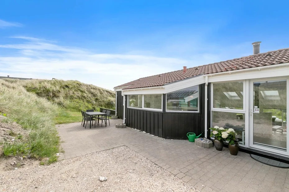 Una casa baja con patio pavimentado y asientos al aire libre junto a dunas cubiertas de hierba.