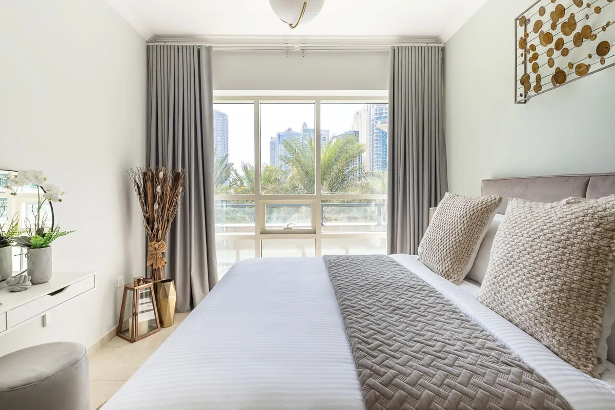 Un dormitorio luminoso con una cama tendida junto a una gran ventana con cortinas.