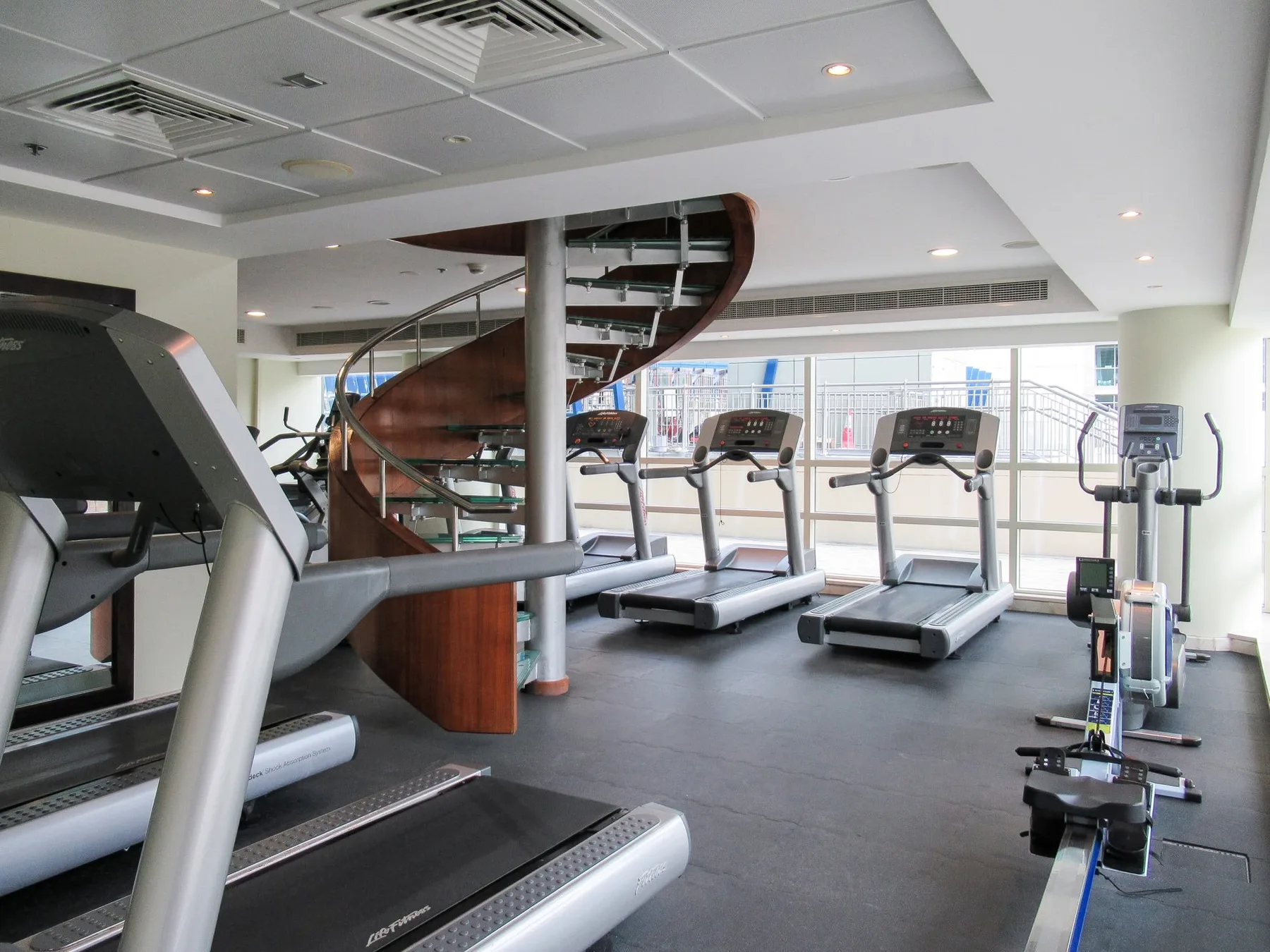Un gimnasio con cintas de correr y equipo de ejercicio junto a grandes ventanas.
