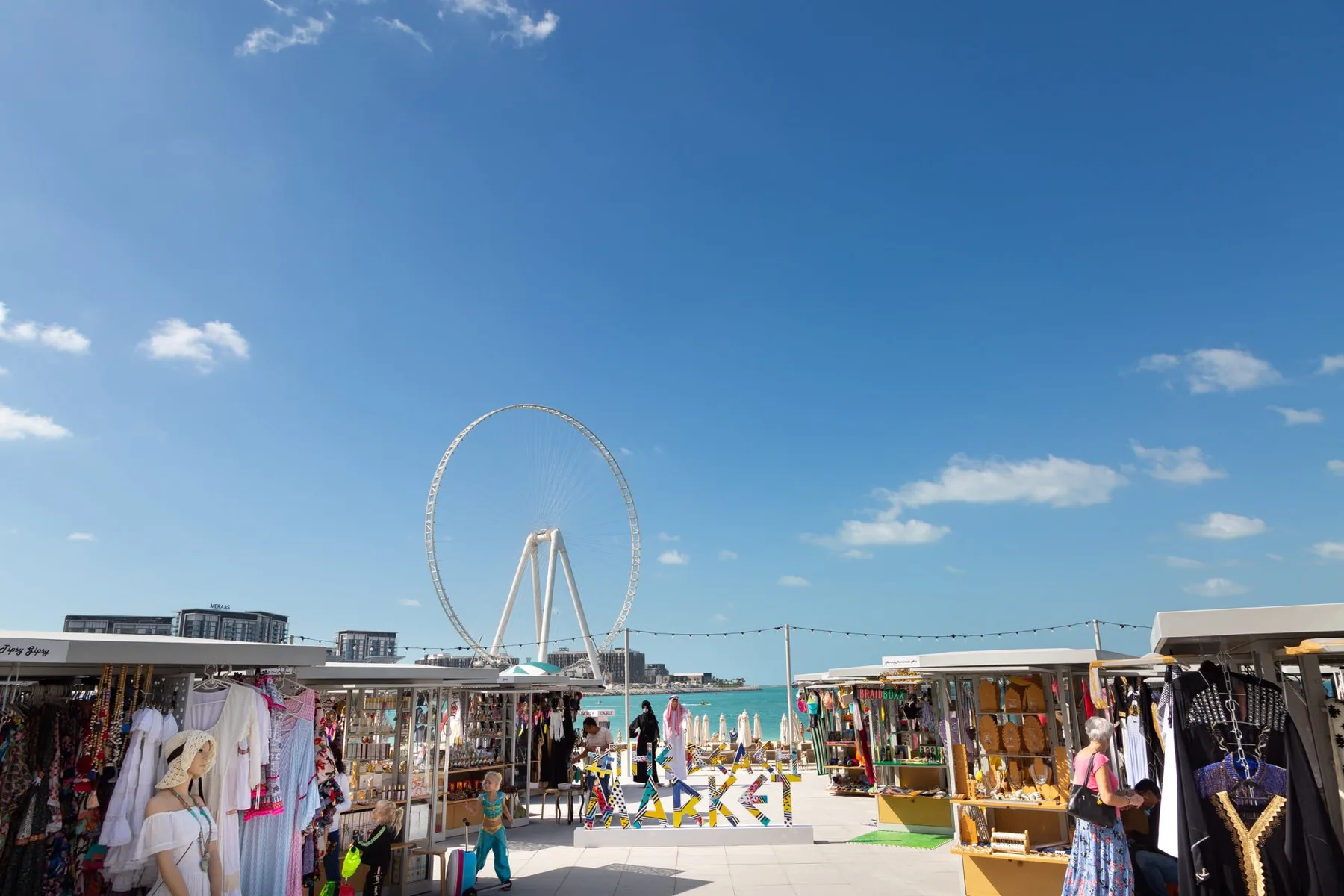 Un mercado junto al mar con puestos de ropa y una gran rueda de observación al fondo.