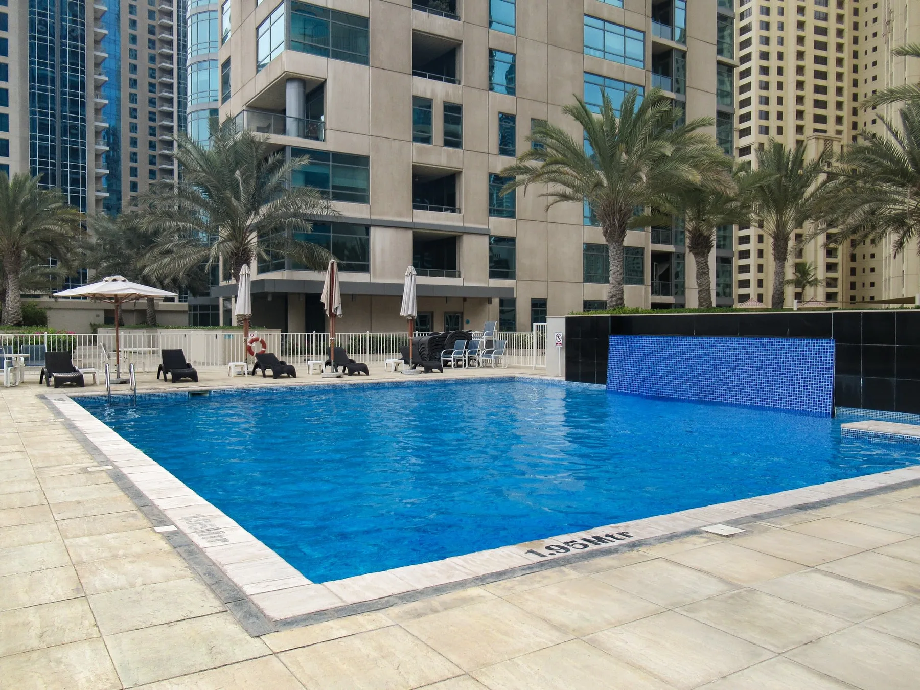 Una piscina está junto a una terraza pavimentada, con palmeras y edificios altos al fondo.