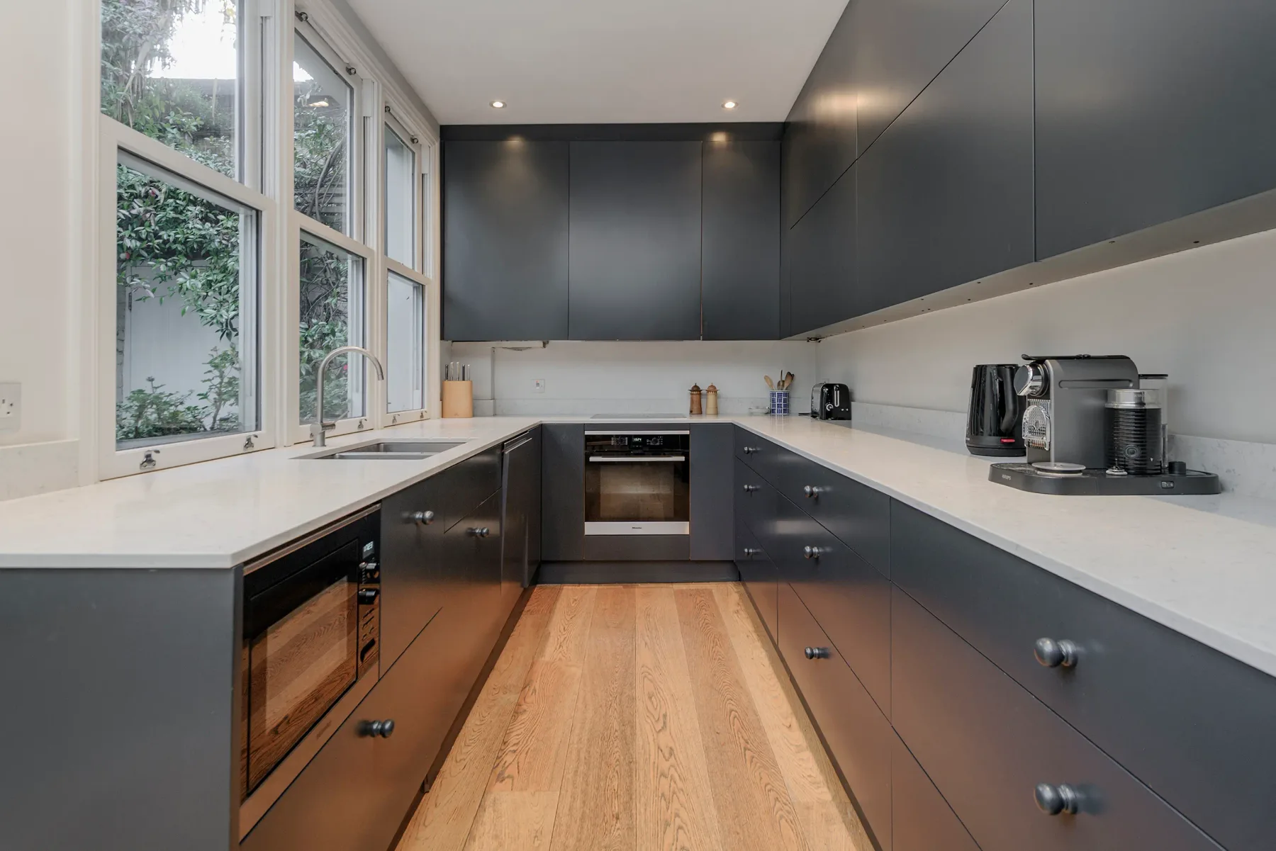 A modern kitchen with dark cabinets, light countertops, and large windows.