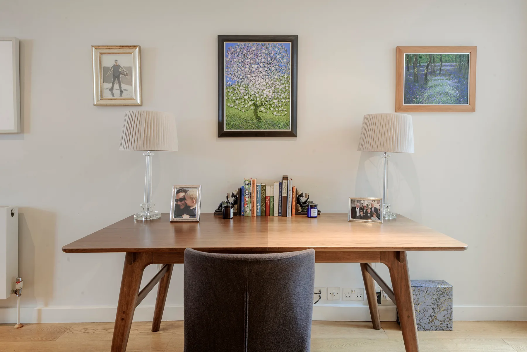A wooden desk with a chair, books, lamps, and framed pictures against a light wall.