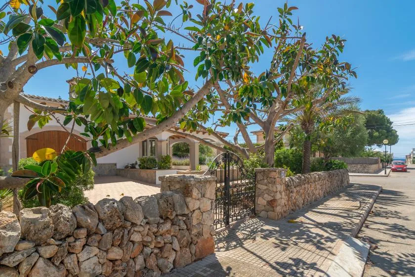 A house entrance with a stone wall and leafy trees beside the road.