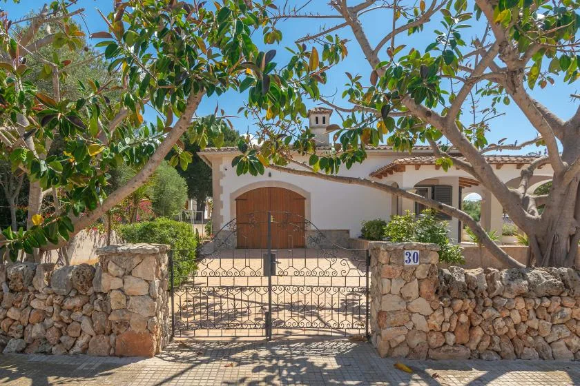 A white house stands behind a metal gate and stone walls, shaded by trees.