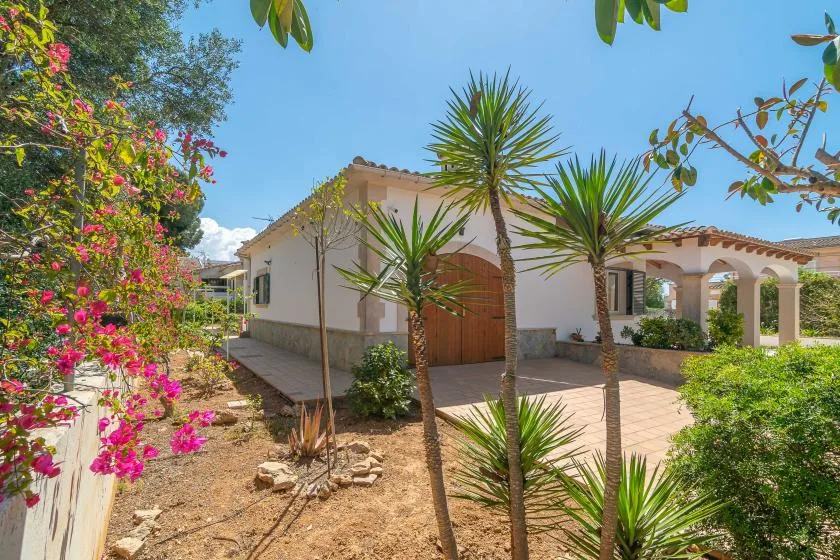 A single-story house with a tiled roof stands beside a paved driveway and flowering plants.