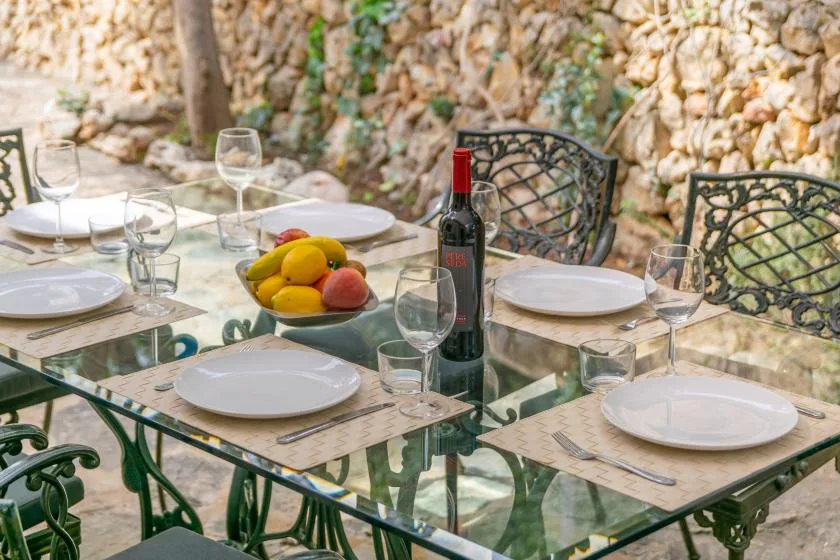 An outdoor dining table is set with plates, glasses, and a bowl of fruit.