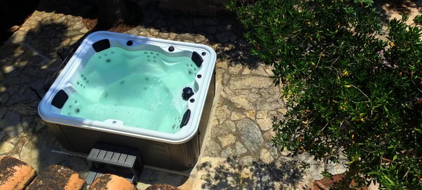 An outdoor hot tub sits beside a stone-paved area and greenery.