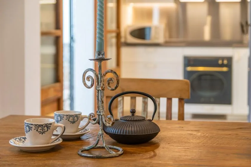 A wooden dining table with teacups and a kettle sits in front of a kitchen.