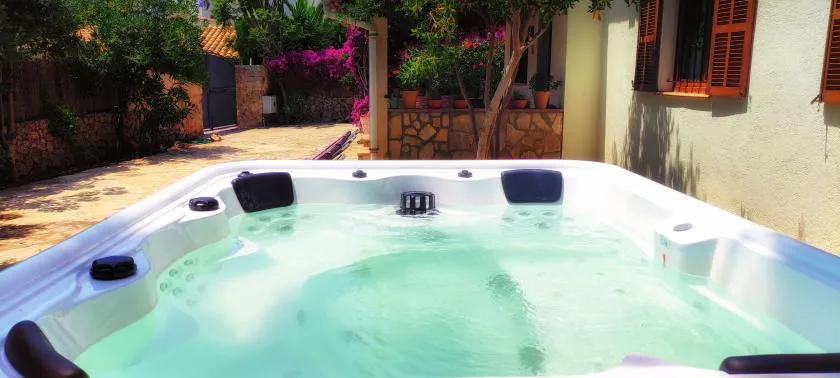 An outdoor hot tub beside a house, surrounded by greenery and flowering plants.
