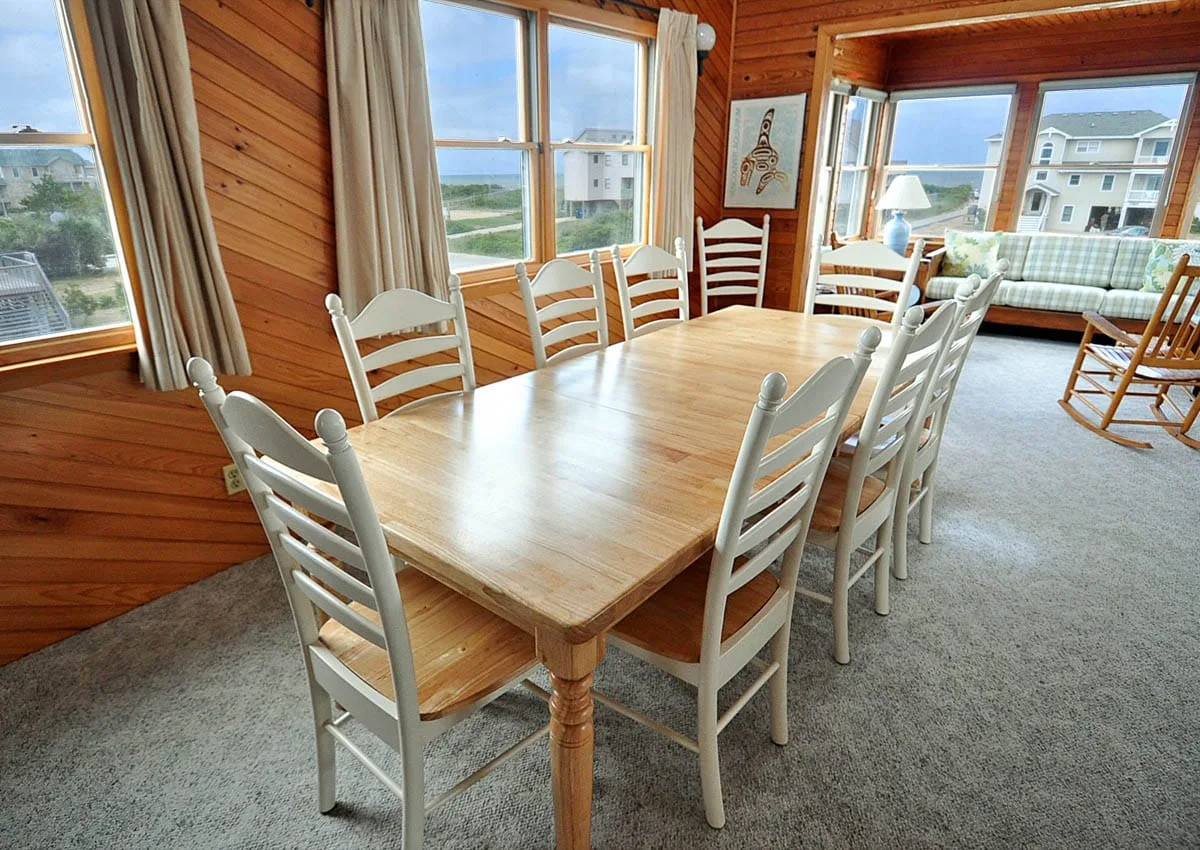 Comedor con una larga mesa de madera y sillas blancas junto a grandes ventanas.