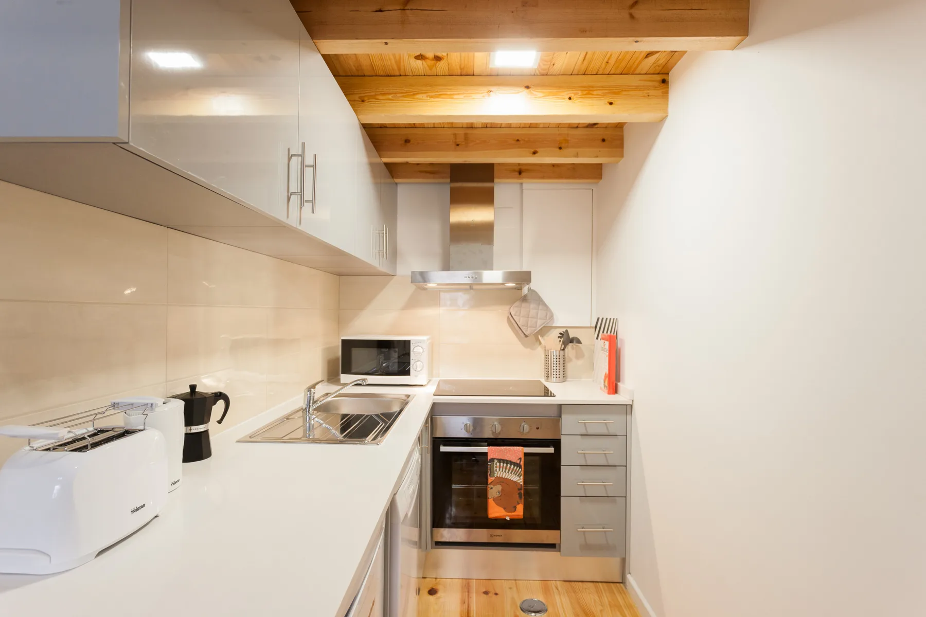 Una cocina compacta con encimeras blancas, electrodomésticos integrados y vigas de madera en el techo.
