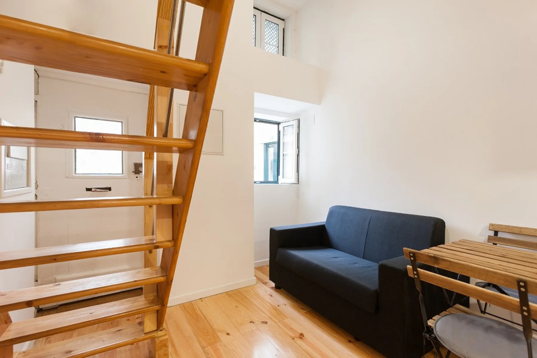 A bright studio living area with dark seating, a wooden table, and stairs.
