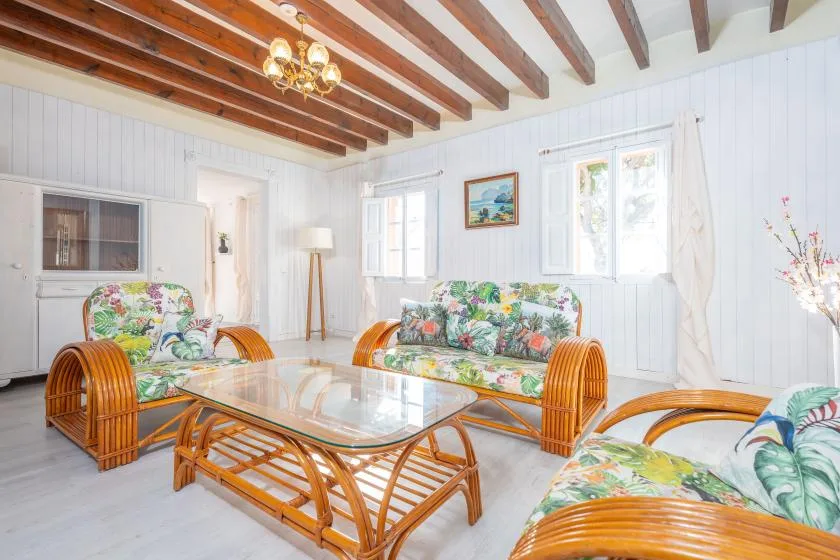 A bright living room with floral seating and glass-topped tables beneath exposed wooden beams.